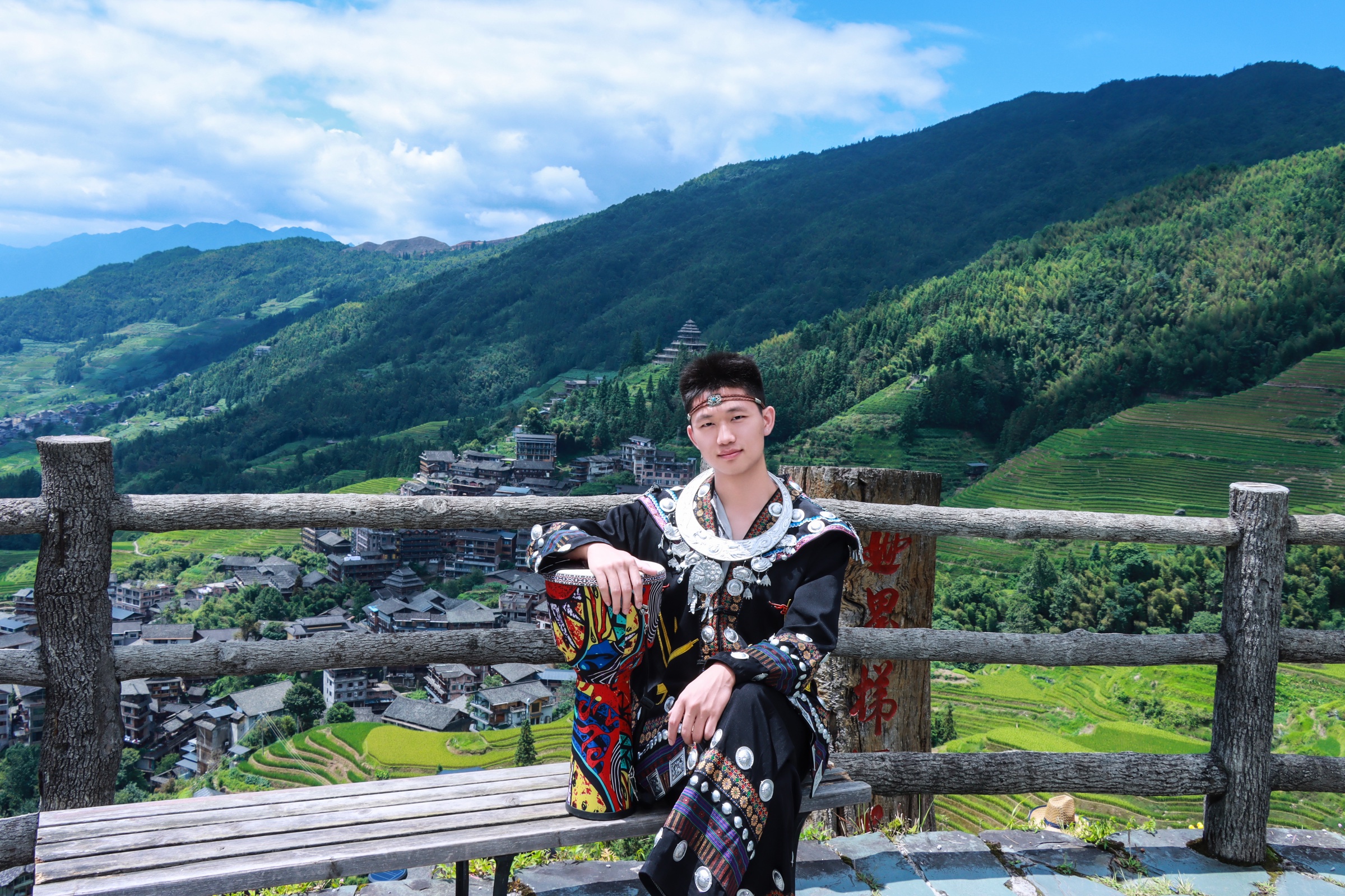 祖仁泽在一次外出旅行中体验民族服装,身后有山林、村落与梯田