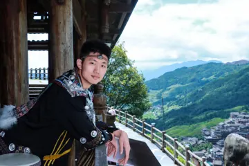 外出旅行体验民族服装，坐在木质栏杆旁，身后是山林与村落