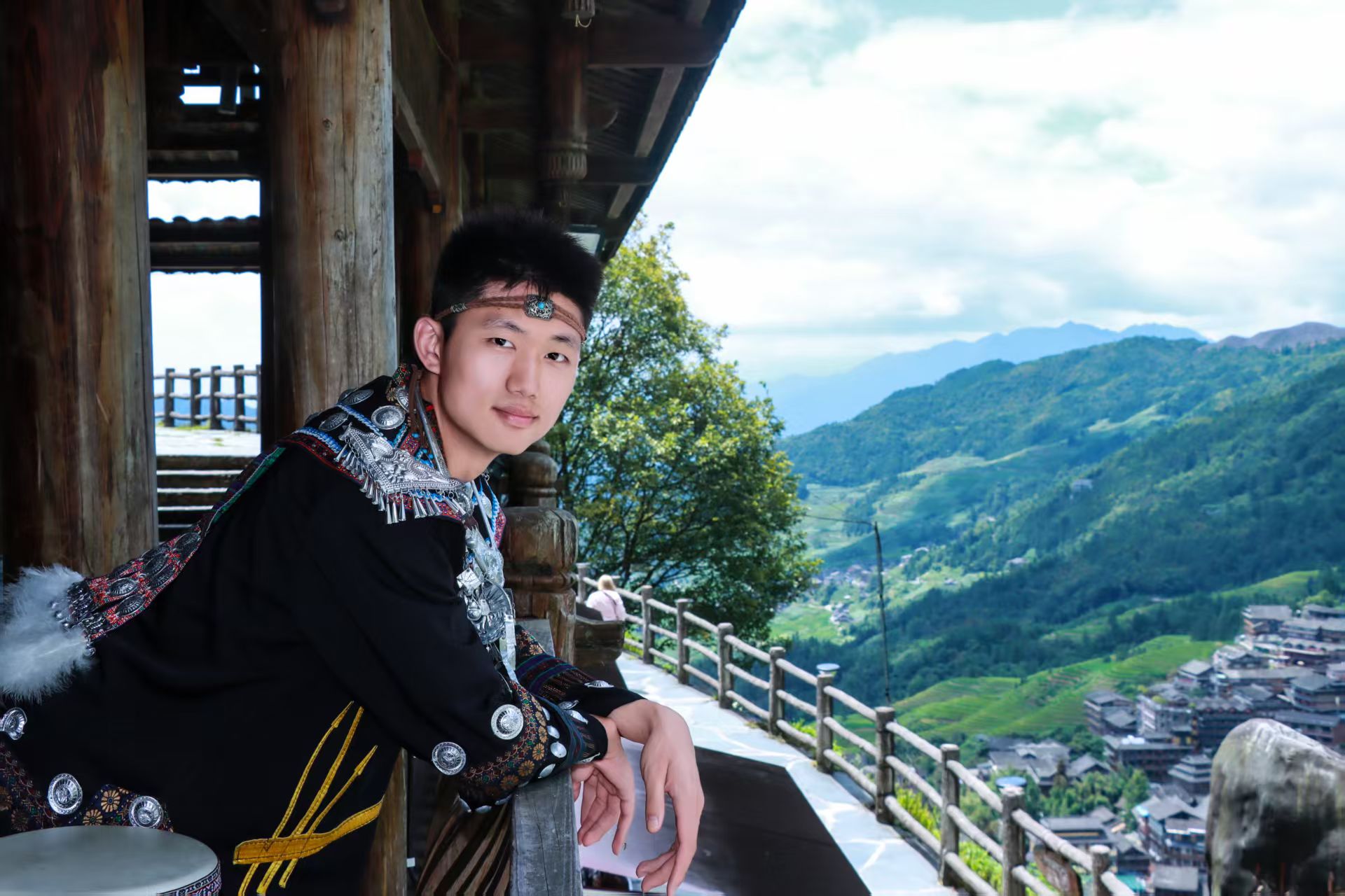 外出旅行体验民族服装,坐在木质栏杆旁,身后是山林与村落